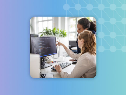 Two people collaborating at a computer as one points to code on a monitor