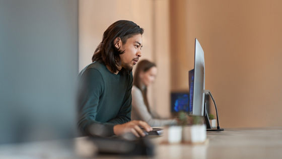Person seated at a desk using a desktop computer in an office, with another person working at a computer in the background