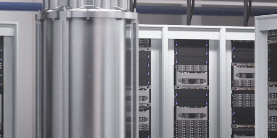 Cryogenic quantum computing hardware housed in a cylindrical chamber, surrounded by multiple server racks in a data center environment.