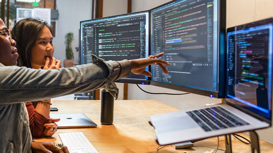 Two people coding together at dual monitors, one pointing at the screen