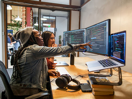 Two people coding together at dual monitors, one pointing at the screen