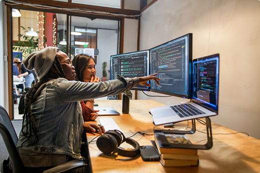 Two people coding together at dual monitors, one pointing at the screen