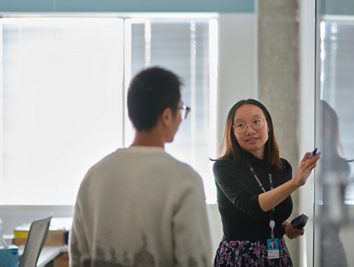 Person pointing at a glass board while another person listens in an office