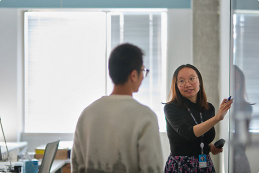 Person pointing at a glass board while another person listens in an office